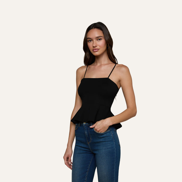 A woman wearing the Parisa Peplum Top—a black sleeveless tank with a fitted bodice—paired with blue jeans stands against a plain light background, facing forward with one hand on her hip and the other relaxed by her side.