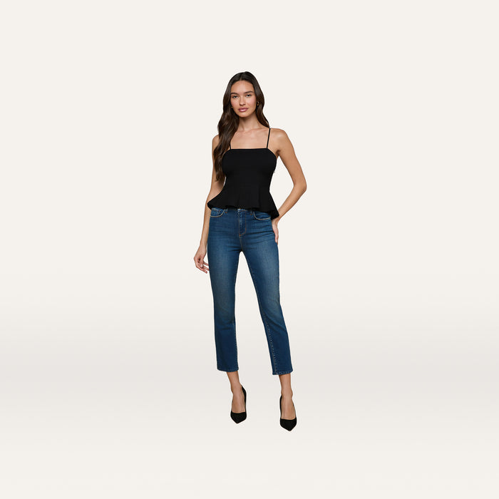 A woman wearing the Parisa Peplum Top—a black tank with adjustable straps—paired with blue jeans and black heels, stands against a plain white background.