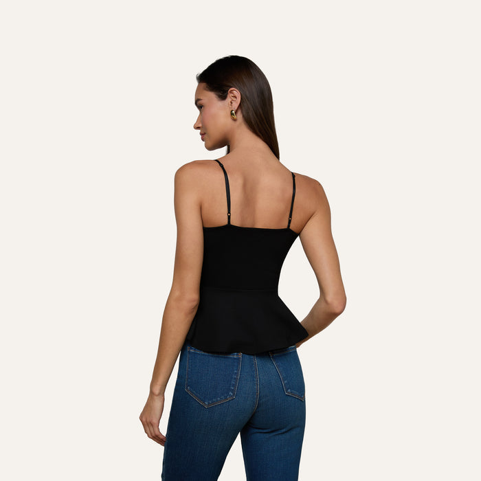 A woman with long brown hair, standing with her back to the camera against a plain light background, wears the Parisa Peplum Top—a black tank featuring a fitted bodice—paired with blue jeans.