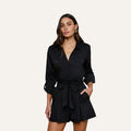 The woman is wearing the Pearson Poplin Shirt in black, styled with a belted waist and buttoned chest pockets, resembling a romper with long sleeves, as she stands with one hand in her pocket against a plain light background.
