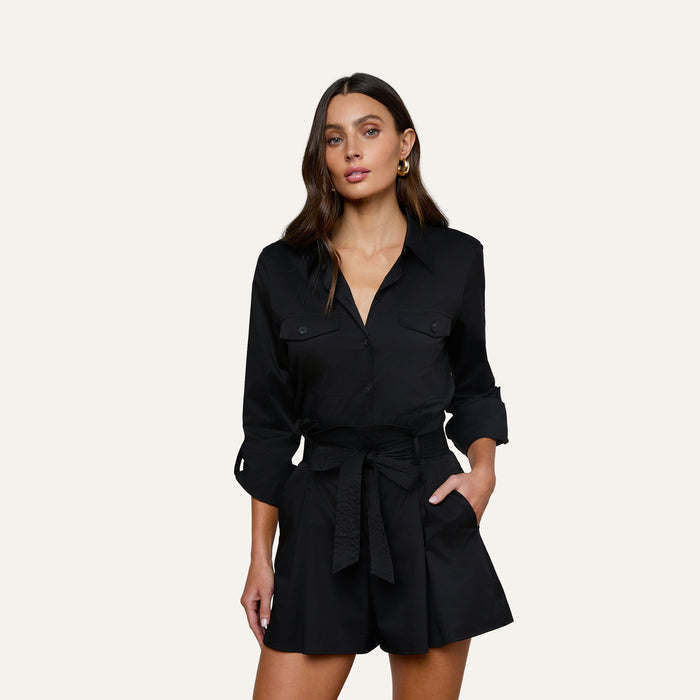 The woman is wearing the Pearson Poplin Shirt in black, styled with a belted waist and buttoned chest pockets, resembling a romper with long sleeves, as she stands with one hand in her pocket against a plain light background.