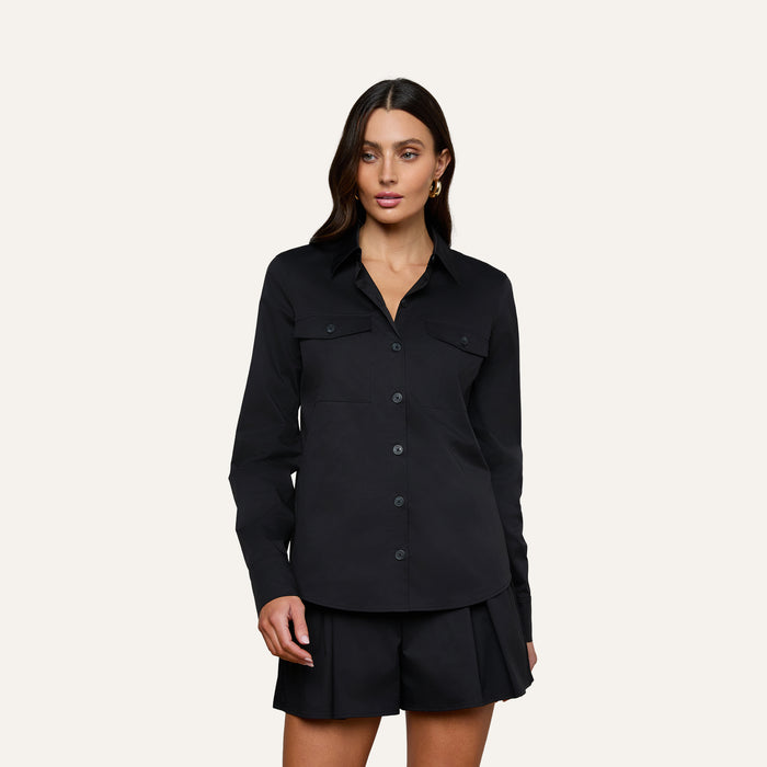 A woman stands against a plain background wearing the Pearson Poplin Shirt in black, featuring two chest pockets and matching black shorts.