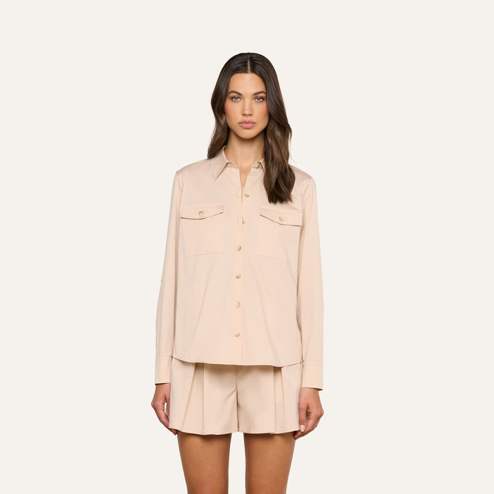 A woman wearing the Pearson Poplin Shirt in light beige, featuring long sleeves and front pockets, paired with matching pleated shorts, stands against a plain white background.