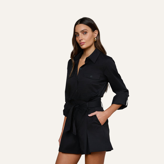 A woman wearing the Pearson Poplin Shirt, a black button-down dress with rolled-up long sleeves, stands with one hand in her pocket against a plain, light background.