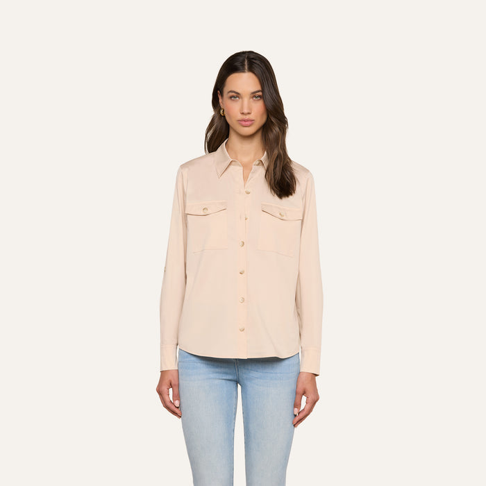 A woman stands against a plain background, wearing the Pearson Poplin Shirt in light beige—an utility-inspired button-down with front pockets—paired with light blue jeans. Her long brown hair is styled in loose waves, and she has a neutral facial expression.
