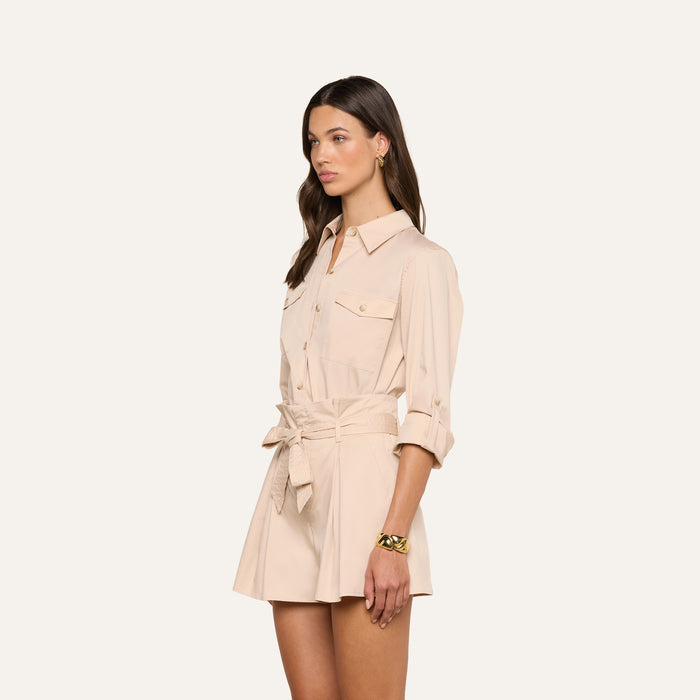 A woman models the Pearson Poplin Shirt, a light beige, utility-inspired collared shirt dress featuring rolled sleeves, front pockets, and a tied waist belt, standing against a plain white background.