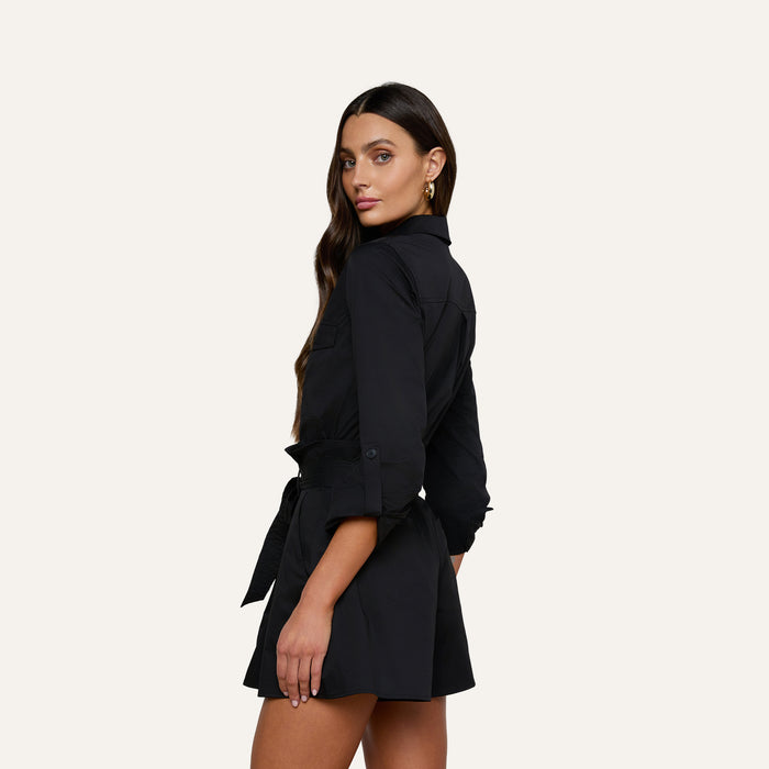 A woman wearing the Pearson Poplin Shirt, styled with long sleeves and a belted waist to resemble a button-down romper, stands slightly turned to the side, looking at the camera against a plain light background.