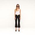 A woman stands against a plain white background, wearing black sunglasses, a white tank top, and the Petite Kendra Coated Jean—a pair of high-rise black cropped pants with a gold-buckle belt—paired with black heels.