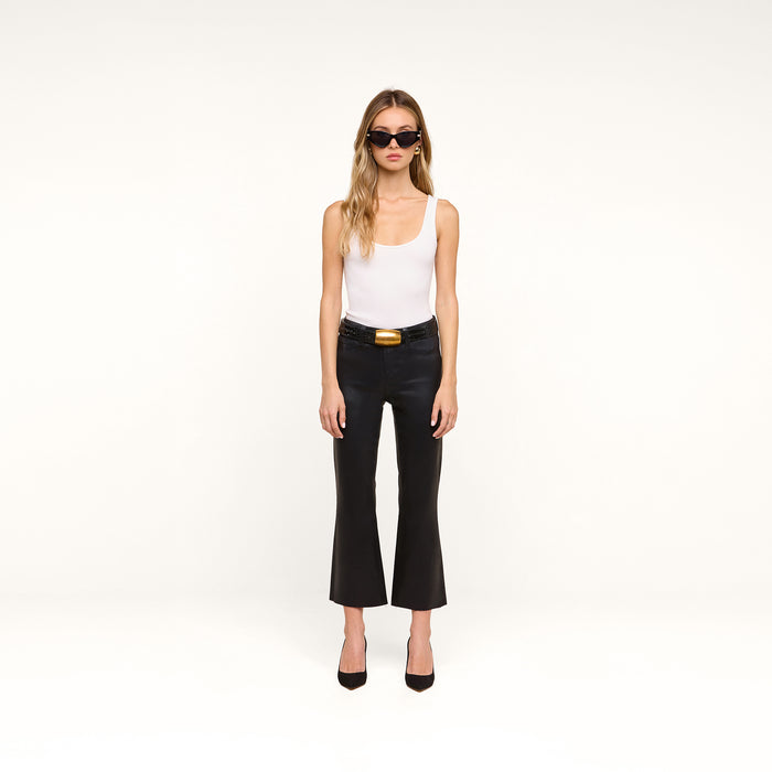 A woman stands against a plain white background, wearing black sunglasses, a white tank top, and the Petite Kendra Coated Jean—a pair of high-rise black cropped pants with a gold-buckle belt—paired with black heels.