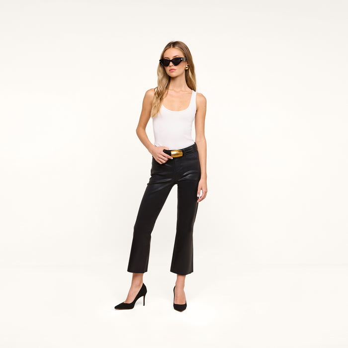 A woman wearing sunglasses, a white tank top, black Petite Kendra Coated Jeans, and black heels stands against a plain white background.