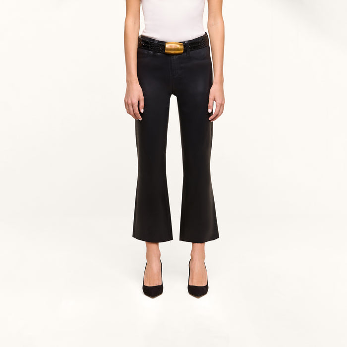 Person wearing a white sleeveless top and the Petite Kendra Coated Jean—black high-rise flared pants styled with a wide black belt featuring a gold buckle—and black pointed-toe heels, standing against a plain white background.