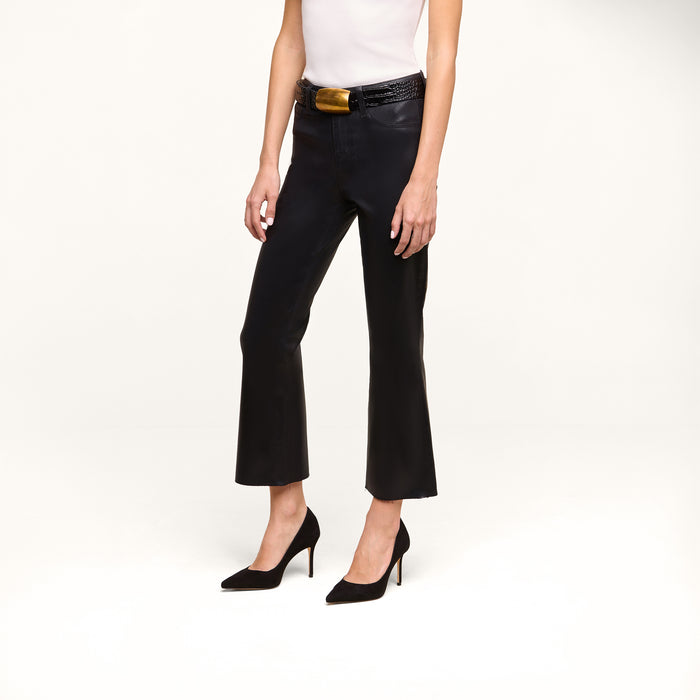 Wearing the Petite Kendra Coated Jean, a person stands against a plain white background, styled with a white sleeveless top, black belt with a gold buckle, and black pointed-toe heels.