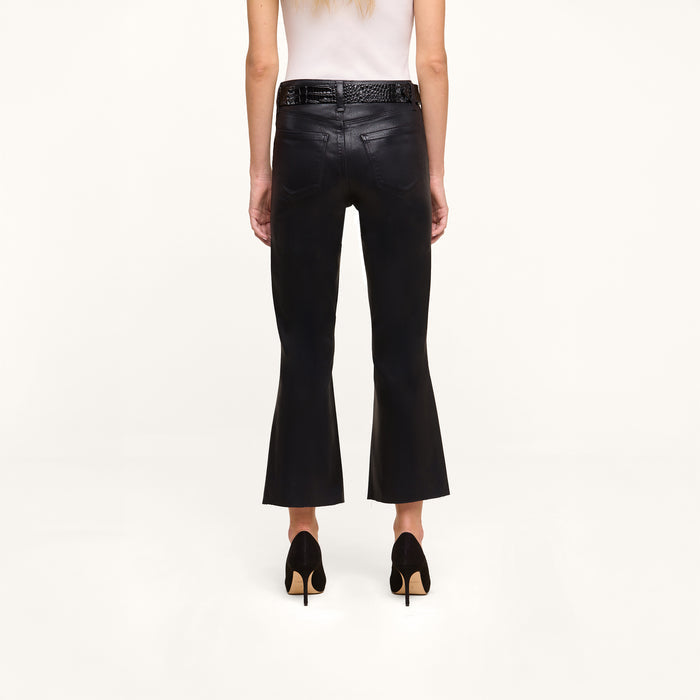 A person stands facing away against a plain white background, wearing the Petite Kendra Coated Jean in black high rise denim, paired with a white sleeveless top, black high heels, and a black braided belt.