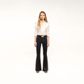 A woman stands against a white background, wearing a white button-up blouse, the Petite Marty Coated Jean in black high-rise petite flare style, a black belt with a gold buckle, and black heeled sandals.