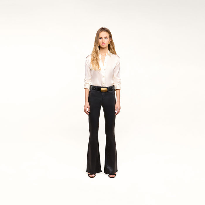 A woman stands against a white background, wearing a white button-up blouse, the Petite Marty Coated Jean in black high-rise petite flare style, a black belt with a gold buckle, and black heeled sandals.