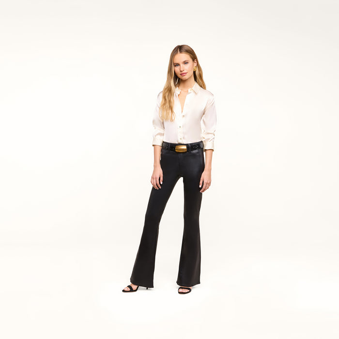A woman stands against a plain white background wearing a white button-up blouse and the Petite Marty Coated Jean—a pair of black high-rise coated denim flared pants—styled with black heeled sandals and a black belt with a large gold buckle.