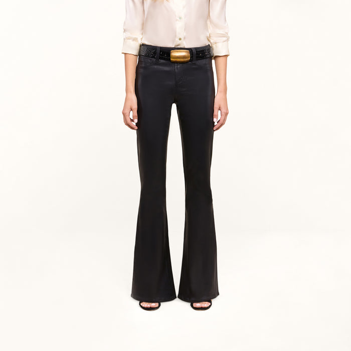 Person wearing a cream blouse, the Petite Marty Coated Jean in black high rise flare style, a black belt with a large gold buckle, and black open-toe heels, standing against a plain light background. Upper face is not visible in the image.