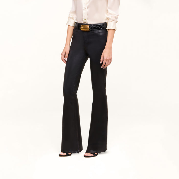 A person wearing a cream blouse, black belt, the Petite Marty Coated Jean in high rise petite flare style, and black heeled sandals stands against a plain white background.