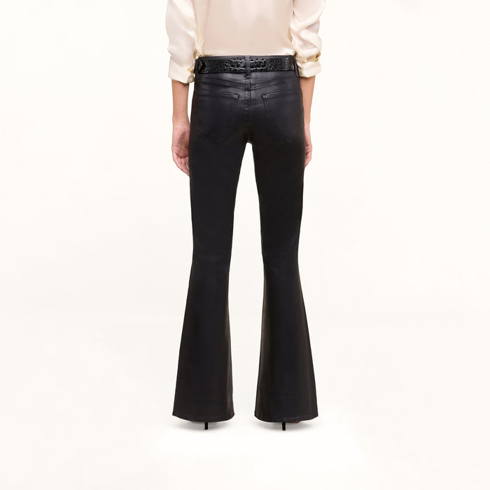 A person stands facing away, wearing the Petite Marty Coated Jean—high-rise black flared pants—paired with a cream long-sleeve shirt tucked in and black high-heeled shoes. The background is plain white.
