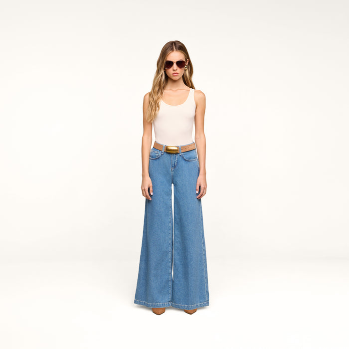 A person wearing sunglasses, a beige sleeveless top, the Petite Quincy Wide-Leg Jean in blue with a brown belt and brown shoes stands against a plain white background.