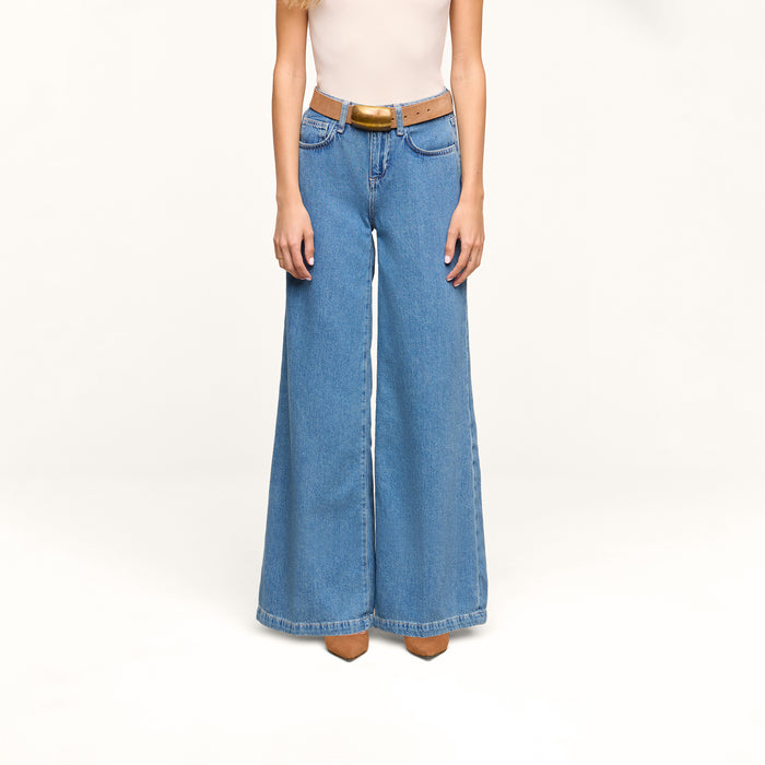 A person wearing the Petite Quincy Wide-Leg Jean in featherweight denim, a tan belt, a light-colored sleeveless top, and pointed brown shoes stands against a plain white background.