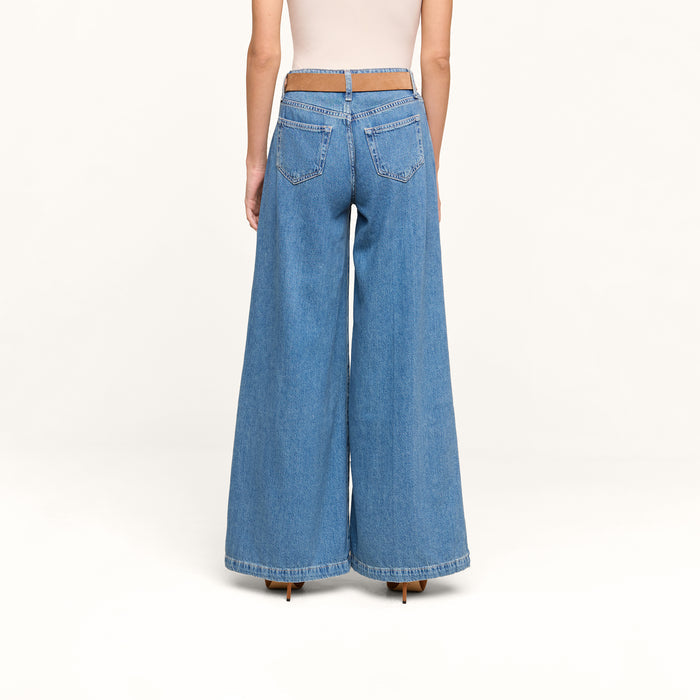 A person stands facing away, wearing the Petite Quincy Wide-Leg Jean in featherweight blue denim with a cut-out waistband and a beige top, set against a plain, light background.