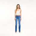 A woman stands against a plain white background wearing a white tank top, brown belt, brown shoes, and Petite Ruth Straight-Leg Jean in heritage indigo wash.