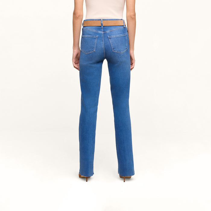 A person stands facing away, wearing the Petite Ruth Straight-Leg Jean in a heritage indigo wash featuring a cut-out section at the waistband, paired with a beige top. The background is plain white.