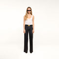 A woman with long blonde hair wears black sunglasses, a white tank top, and the Petite Scottie Coated Jean with a belt and black shoes, standing against a plain white background.