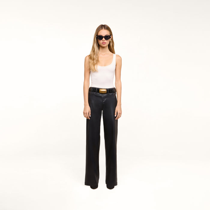 A woman with long blonde hair wears black sunglasses, a white tank top, and the Petite Scottie Coated Jean with a belt and black shoes, standing against a plain white background.