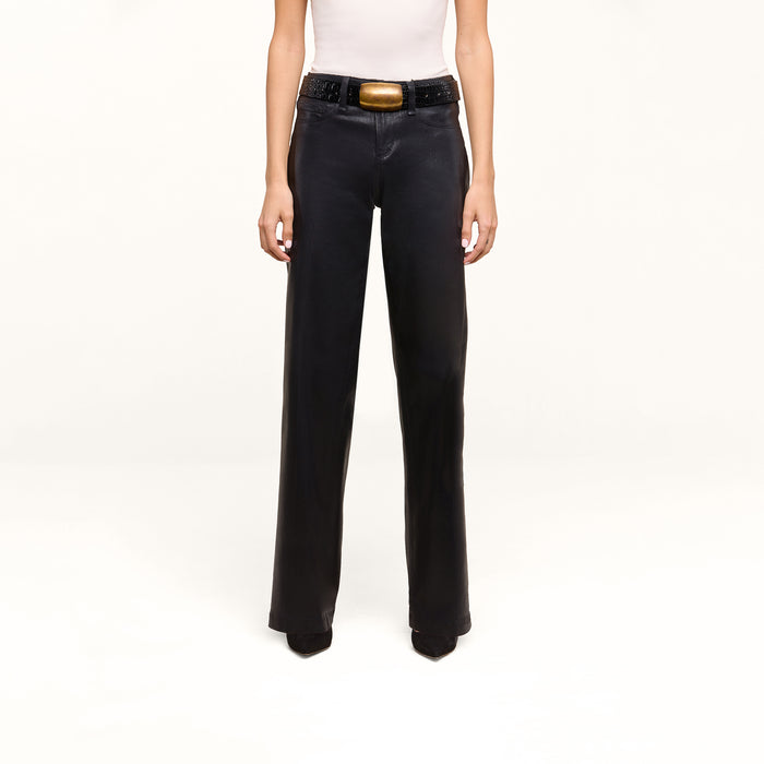 A person is wearing the Petite Scottie Coated Jean, styled in a high rise black wide-leg silhouette, paired with a white sleeveless top and accessorized with a black belt featuring a large gold buckle, standing against a plain white background.