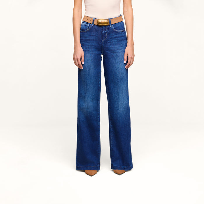 A person stands against a plain white background wearing the Petite Scottie Wide-Leg Jean in blue, paired with a tan belt, beige sleeveless top, and brown pointed shoes.