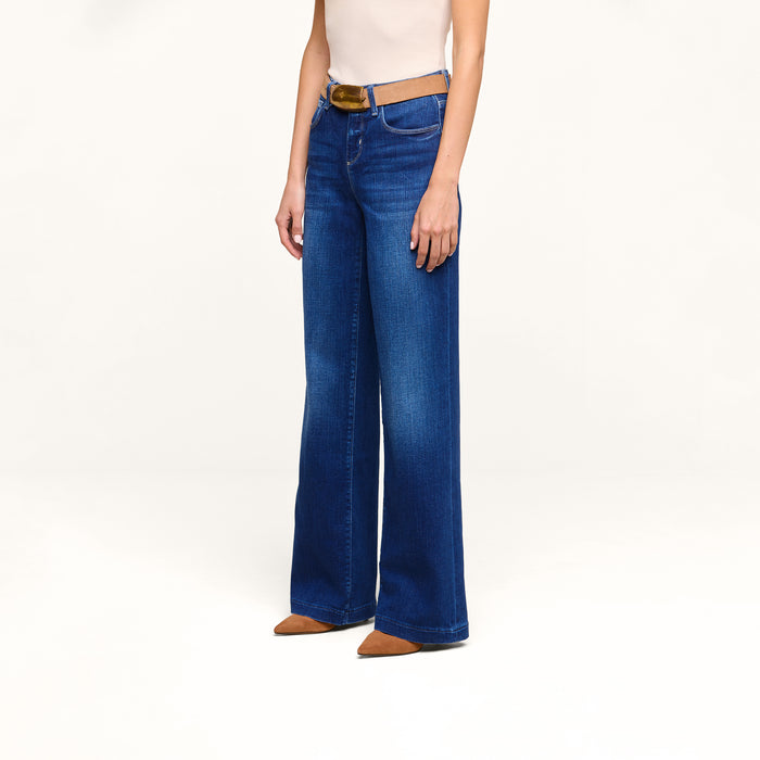 A person is wearing the Petite Scottie Wide-Leg Jean in blue with a tan belt, paired with a sleeveless beige top and brown pointed-toe shoes, standing against a plain white background.