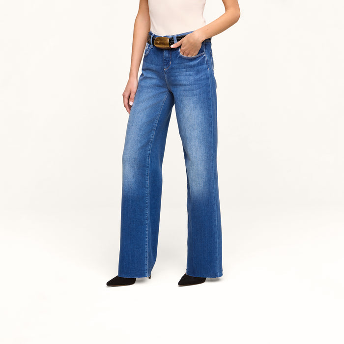 A person is wearing the Petite Scottie Wide-Leg Jean, styled high-rise in blue denim with a brown belt, paired with a white sleeveless top and black pointed-toe shoes, standing against a plain white background.