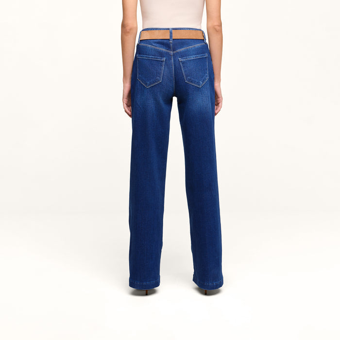 A person is standing facing away, wearing the Petite Scottie Wide-Leg Jean in blue, featuring back pockets and crafted from stretch denim, paired with a light-colored top against a plain white background.