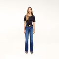 A woman with long blonde hair wears a black t-shirt, the Petite Selma Bootcut Jean in blue super stretch denim, a black belt with a gold buckle, and black sandals, standing against a plain white background.
