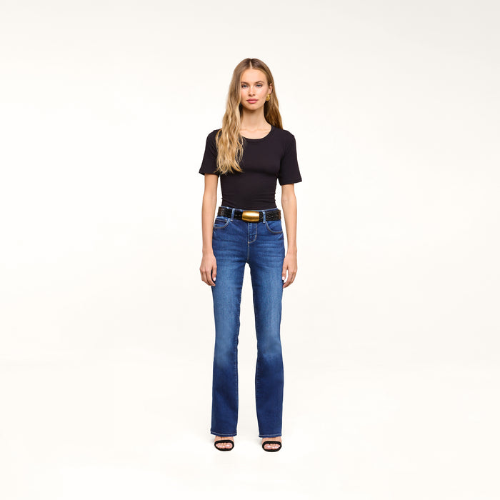 A woman with long blonde hair wears a black t-shirt, the Petite Selma Bootcut Jean in blue super stretch denim, a black belt with a gold buckle, and black sandals, standing against a plain white background.