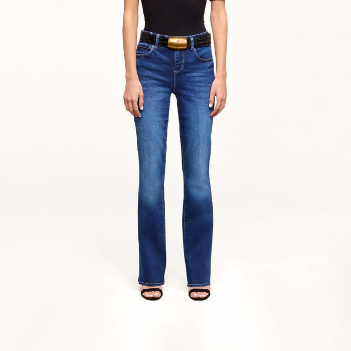 A person wearing a black sleeveless top, the Petite Selma Bootcut Jean in blue super stretch denim, a black belt with a gold buckle, and black heeled sandals stands against a plain white background.