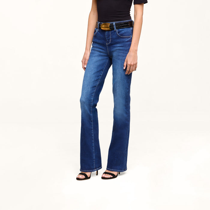 A person wearing a black short-sleeve top, the Petite Selma Bootcut Jean in blue, a black belt, and black high-heeled sandals stands against a plain white background.
