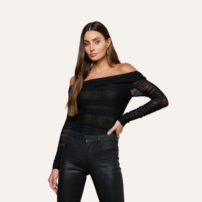 A woman with long brown hair poses with one hand on her hip against a plain light background, wearing the Poppie Off-The-Shoulder Bodysuit in black stretch mesh paired with black pants.