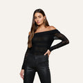 A woman with long brown hair poses with one hand on her hip against a plain light background, wearing the Poppie Off-The-Shoulder Bodysuit in black stretch mesh paired with black pants.