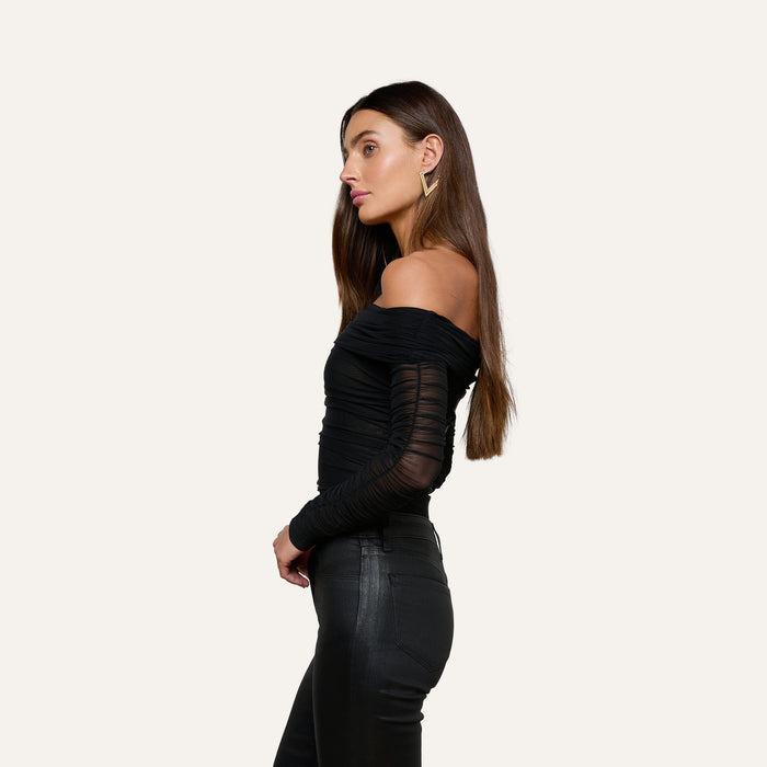 A woman in profile wearing the Poppie Off-The-Shoulder Bodysuit—with stretch mesh sleeves—paired with black fitted pants, standing against a plain, light background.