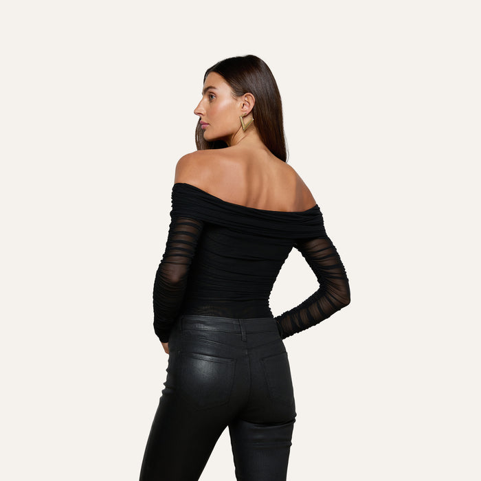 A woman stands facing away, her head turned to the side, wearing the Poppie Off-The-Shoulder Bodysuit paired with black leather pants against a plain light background.