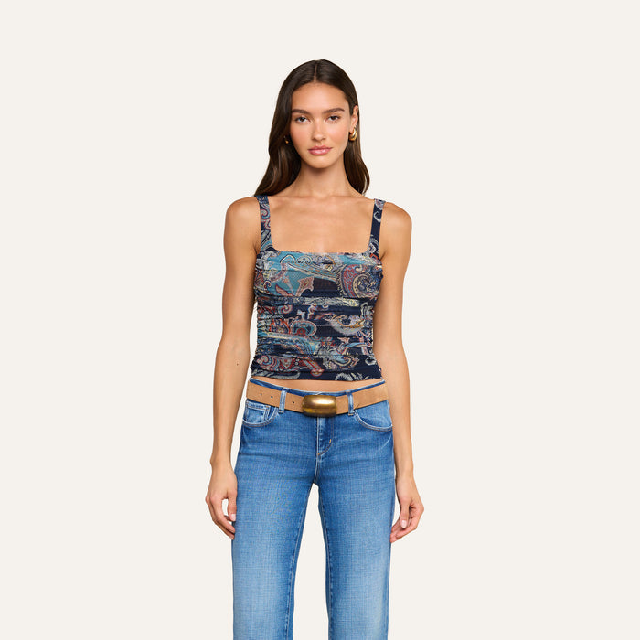 A woman wearing the Rasha Shirred Mesh Tank with a midnight paisley print and square neckline, paired with blue jeans and a tan belt featuring a large buckle, stands against a plain light background, facing forward with a slight smile.