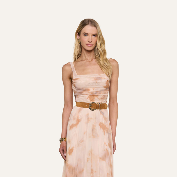 A woman with long blonde hair stands against a plain white background wearing the Rasha Shirred Mesh Tank, a peach and white tie-dye sleeveless top with a square neckline, paired with a tan belt featuring a large circular buckle and accessorized with a gold bracelet.