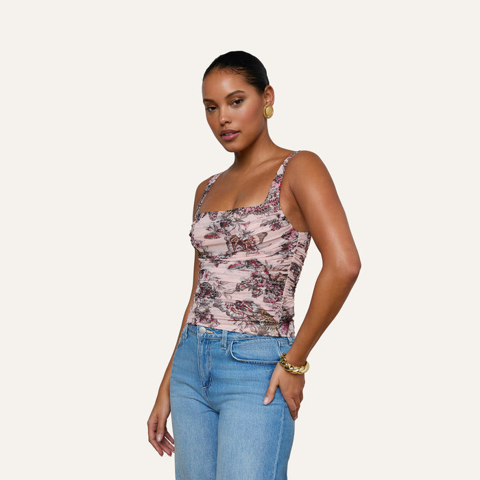 A woman wearing the Rasha Shirred Mesh Tank, which features a square neckline and a butterfly-jungle print, paired with blue jeans, stands with one hand in her pocket against a plain light background.