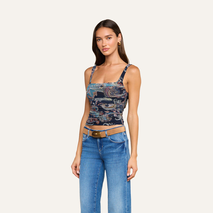 A woman with long brown hair wears the Rasha Shirred Mesh Tank, which features a square neckline and a midnight paisley pattern. She pairs it with blue jeans and a brown belt, standing against a plain light background while facing forward with a relaxed expression.