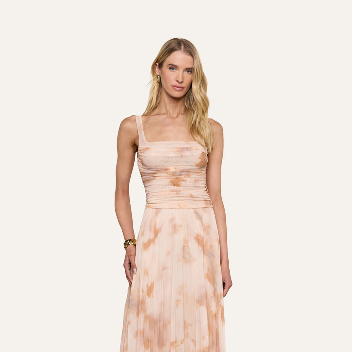A woman with long blonde hair wears the Rasha Shirred Mesh Tank, a sleeveless, square neckline top in a peach and white abstract tie-dye print. She stands facing forward against a plain light background, with a neutral expression and a bracelet on her left wrist.