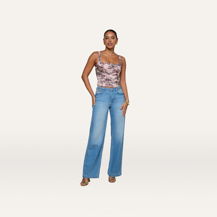 A woman with short hair stands against a plain white background, wearing the Rasha Shirred Mesh Tank—a butterfly-jungle print sleeveless top with a square neckline—paired with light blue wide-leg jeans and heeled sandals. She accessorizes with a bracelet.