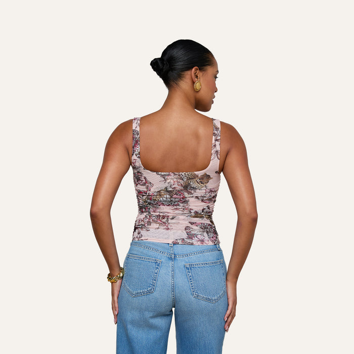 A woman with her hair in a bun is seen from the back, wearing the Rasha Shirred Mesh Tank in a butterfly-jungle print paired with blue jeans, standing against a plain light background.